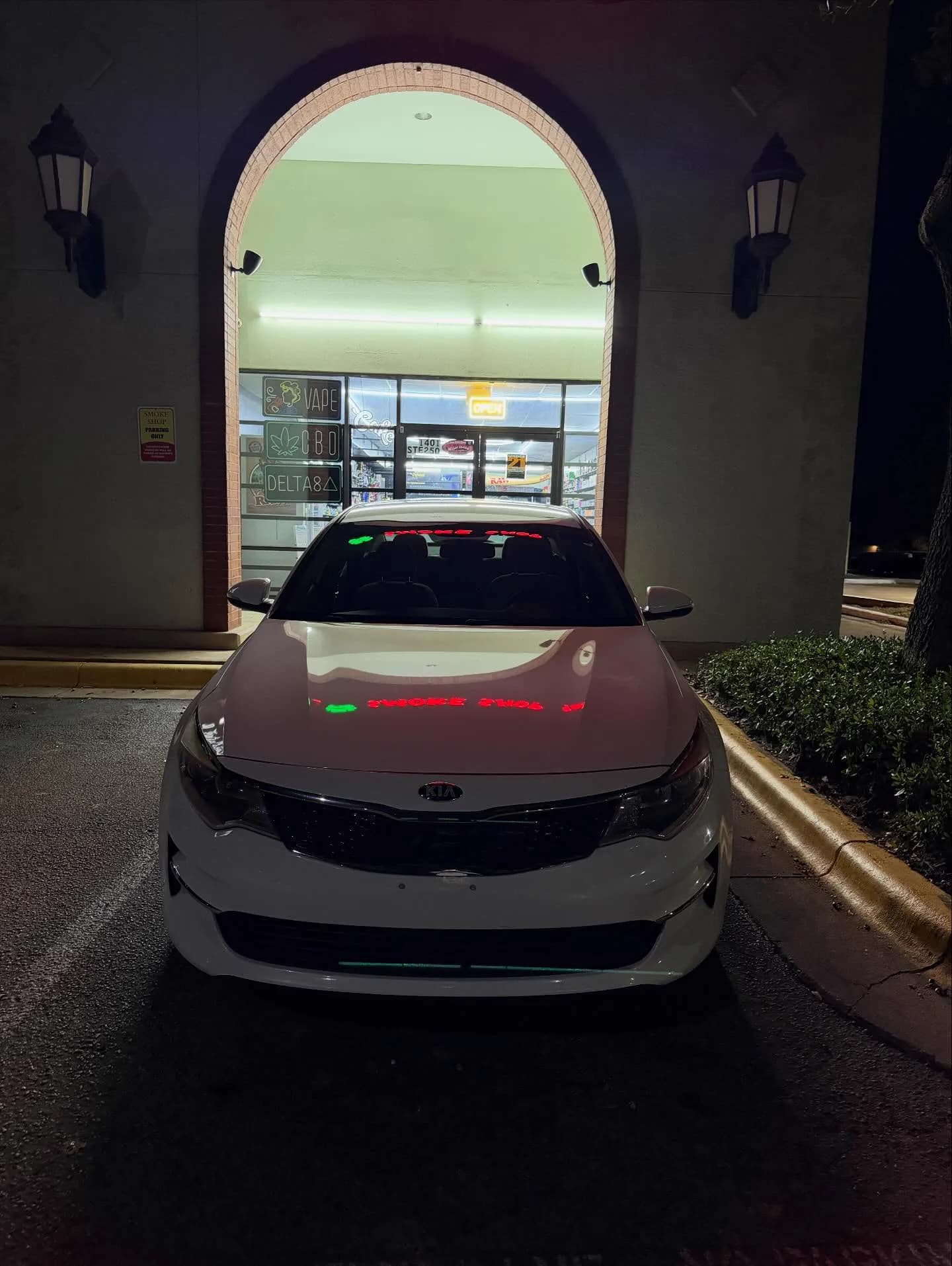 The white Kia Optima photographed head-on at night under a lit archway.